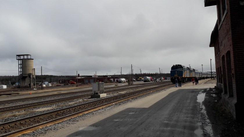 The derelict Hornpayne station on the right; an abandoned coal tower on the left; centre, the Canadian train curving on its tracks into the distance. Grey sky above, grey gravel and concrete below.