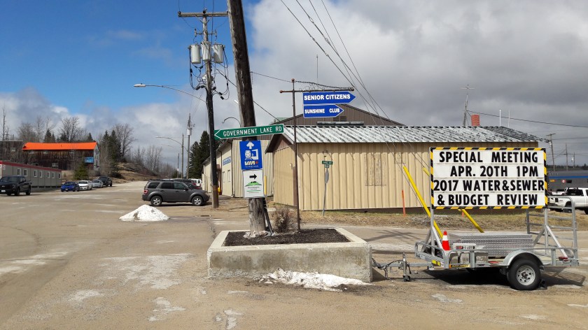 Hornepayne, Ontario: a logging town. Various signs read "Government Lane Rd", "Senior Citizens Sunshine Club" and "Special Meeting Apr. 20th 1pm 2017Water & Sewer & Budget Review". Telegraph poles, dirt road and bare trees, corrugated sheeting huts, clouds and blue in the sky.