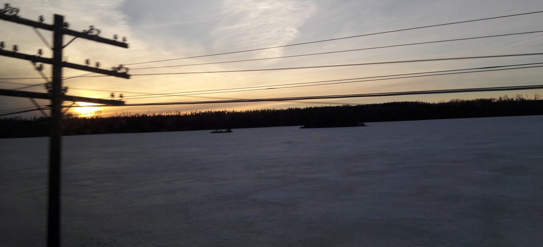 A sunset over an ice-covered lake, black firs in the background and telegraph wires in the foreground.