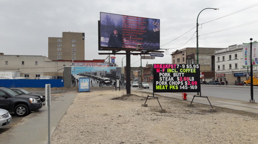 Signs outside Neechi Commons workers' co-op in downtown Winnipeg, with dirt lots and buildings surrounding it. Adverts right and left; central, and artwork with a picture of a human figure and the text: "niidaachag dahapii sinahae / niiya ngiigimoodiimiigo / my body was kidnapped / my spirit lives on"