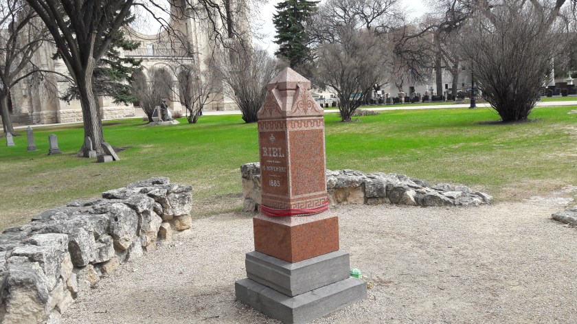 Louis Riel's grave, with a Métis sash wrapped around the column. A low wall surrounds it, and a half-destroyed cathedral is in the background, with a green lawn between.
