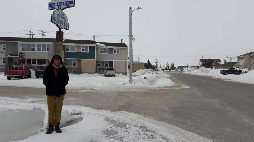 Your correspondent stands beneath a blue street sign reading "Orcade Bay", wrapped up and looking very cold. Snow underfoot, housing blocks behind, and a grey sky.