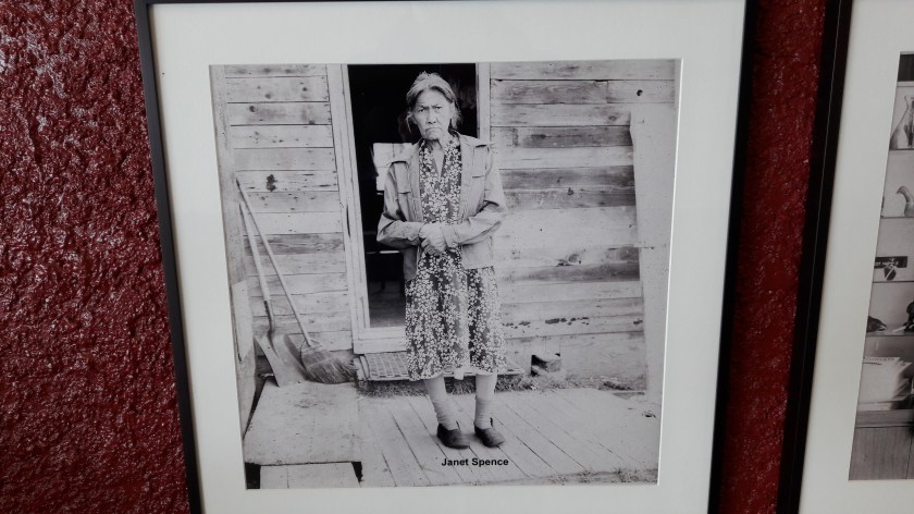 Janet Spence, a late middle aged woman standing in the door of her wooden house. She is wearing a print patterned dress and a short coatel and broom lean in the left of the picture. Her hands are clasped. The photo is labelled at the bottom. The photographer was not labelled in the gallery.