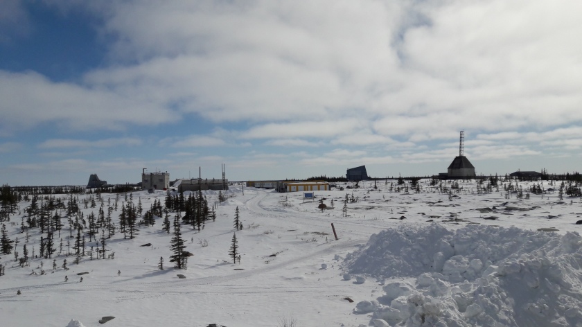 Blue sky and white clouds above, snow drifts and sparse boreal forest below. In between, an old rocket range: blocky huts of different shapes, one with a metal gantry pointing to the sky.