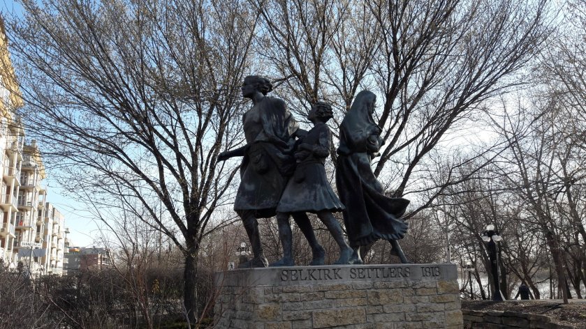 The Selkirk Settler monument, as described in the text. Bare trees behind it; the Red River on the right; buildings and a street on the left.