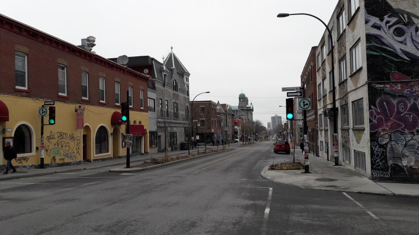 A Montréal street scene with a mixture of new and old architecture, with graffiti and street art visible on the walls and a church far up the street. In the foreground a sign reads "Rue Coloniale".