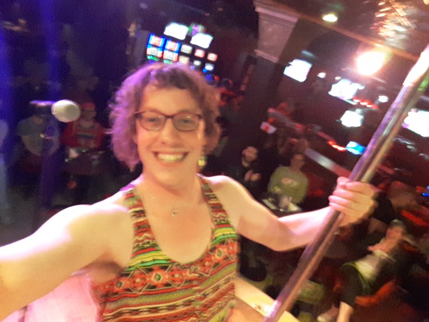 Your correspondent, on stage at the Stock Bar, with a crowd and glowing lights behind. I'm wearing a brightly-patterned strappy top, pink glasses, bouffant hair, and am leaning away from a metal pole.